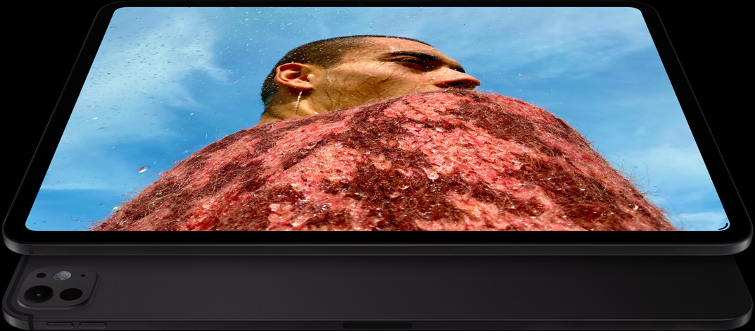 iPad Pro, etupuoli, vaakasuunnassa, tähtimusta väri, pyöristetyt kulmat, musta näytön kehys, näytöllä on punaiseen neuleeseen pukeutunut mies sinisen taivaan alla, alapuolella, toinen iPad Pro, taustapuoli, vaakasuunnassa, tähtimusta väri, ammattitason kamerajärjestelmä, äänenvoimakkuuden lisäyspainike, äänenvoimakkuuden vähennyspainike