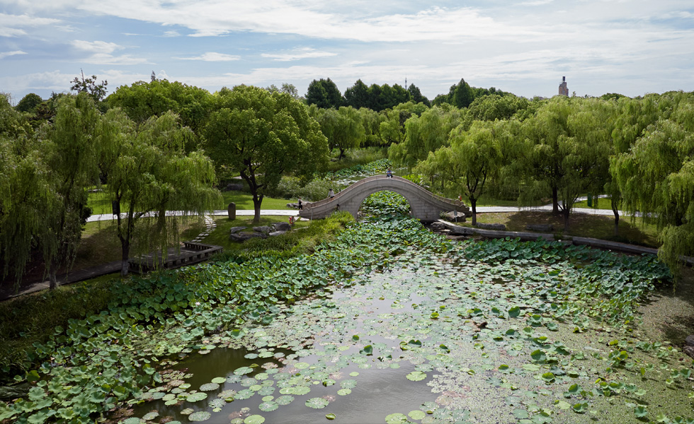 Lush trees surround a pond full of lily pads, and a stone bridge arches over a narrow section of the pond